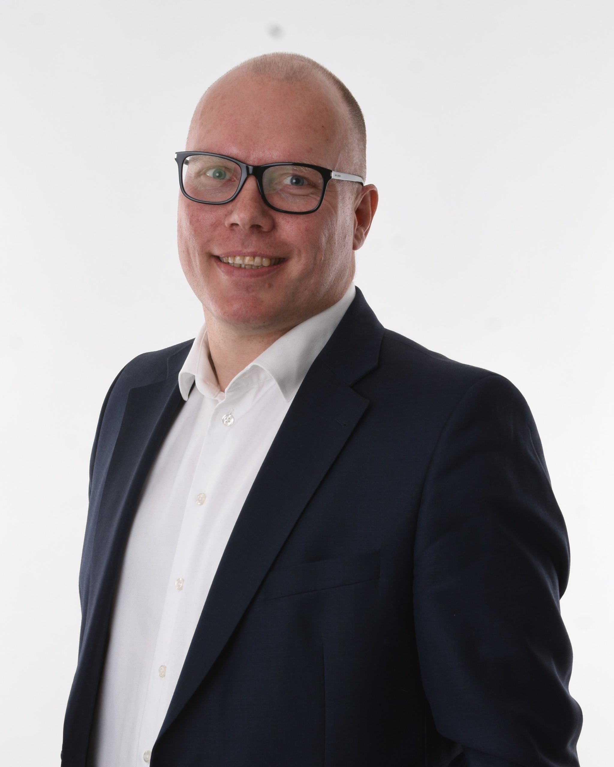 En smilende person iført briller, hvid skjorte og mørk habitjakke poserer på en almindelig hvid baggrund. Peter Nielsen