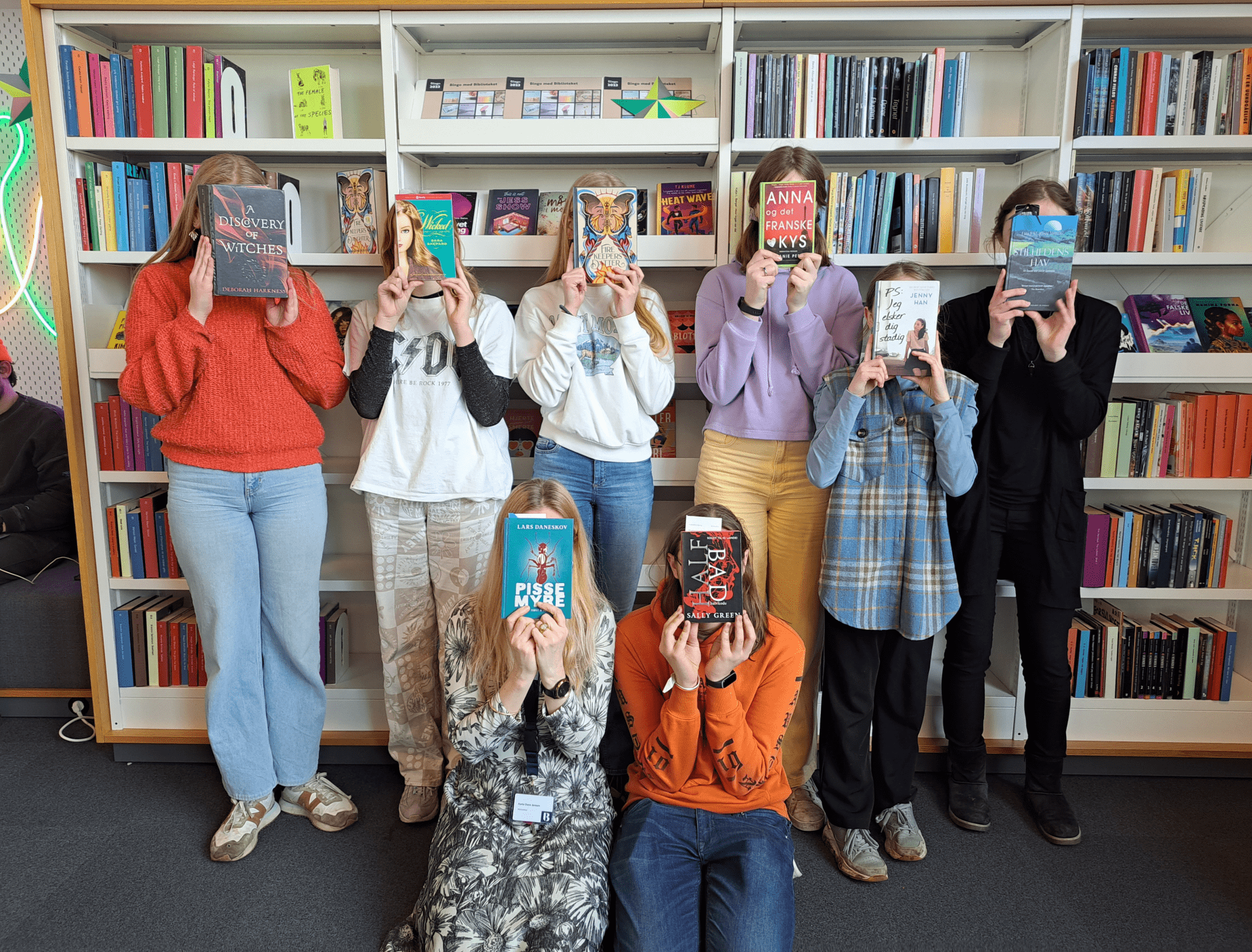 Børn i læseklub holder bøger foran ansigtet i biblioteket.